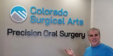 An older man in a blue shirt stands in front of Colorado Surgical Arts' recision oral surgery signage.